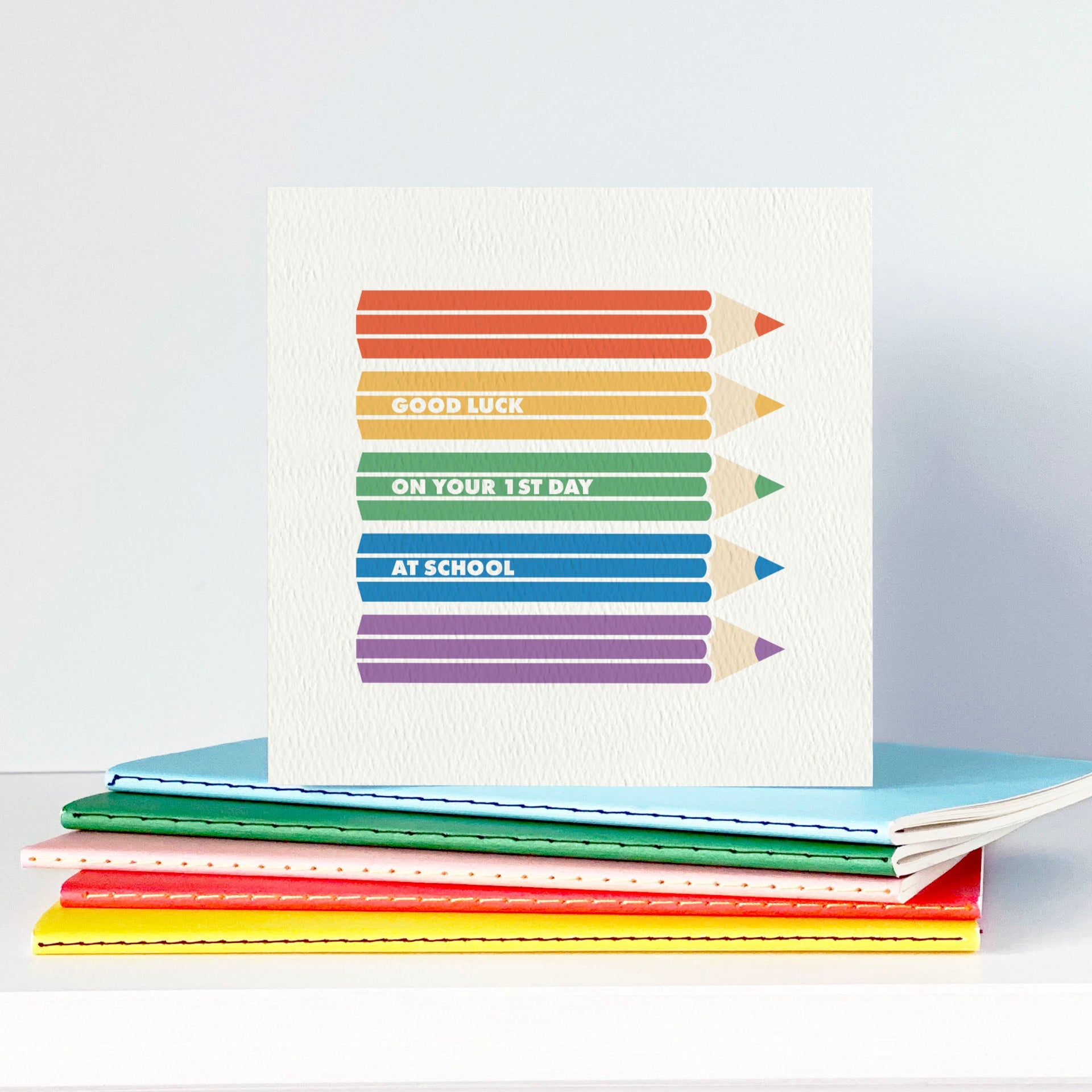 Stack of colourful notebooks with a card featuring rainbow coloured pencils on a white surface.