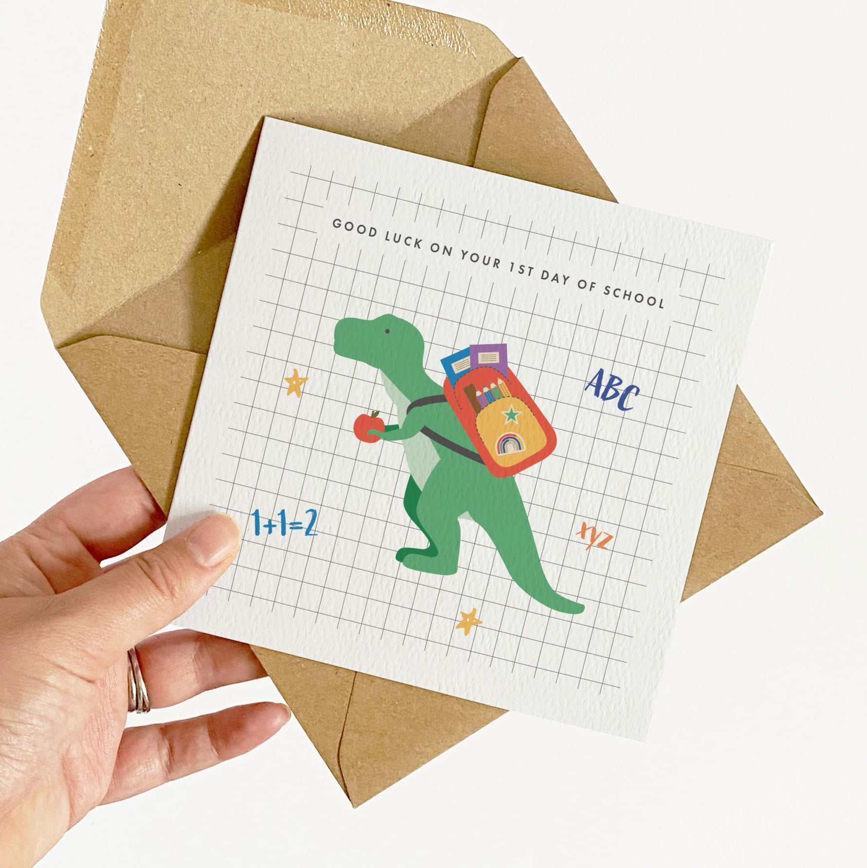 Hand holding a card with a dinosaur illustration and a brown envelope on a white background