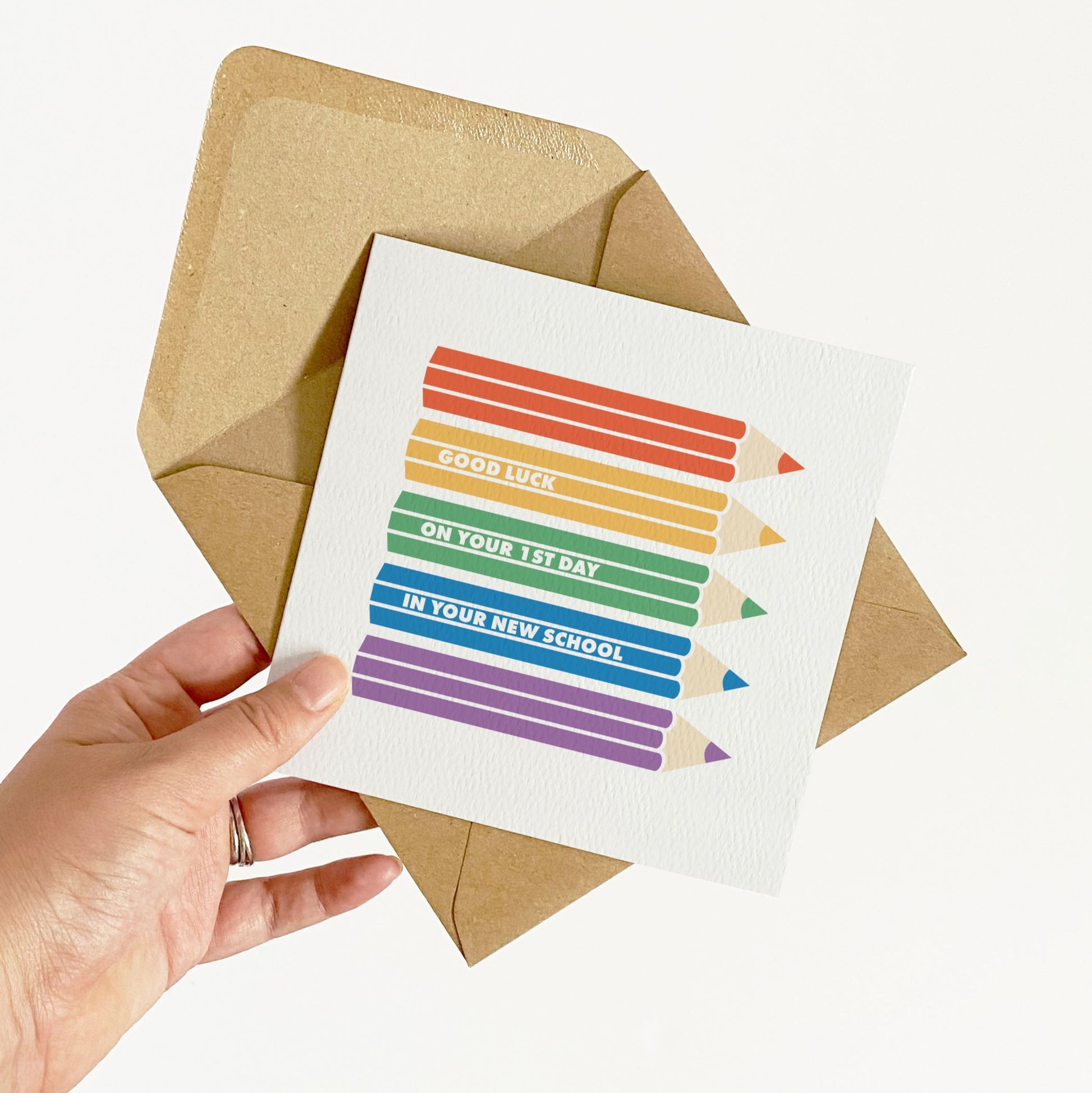 Hand holding a card with rainbow-colored pencils and text, placed on a brown envelope.