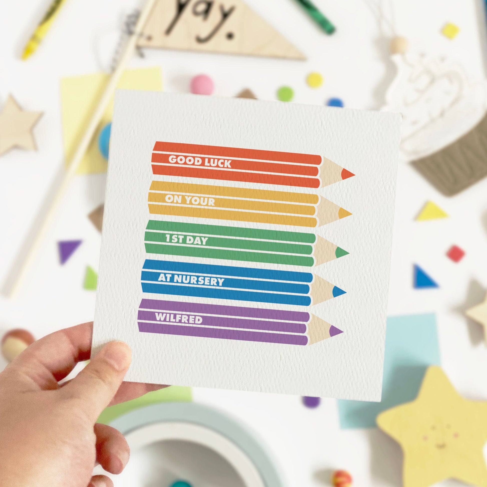 Hand holding a card with rainbow-colored pencils and text, surrounded by colorful stationery items on a white background.