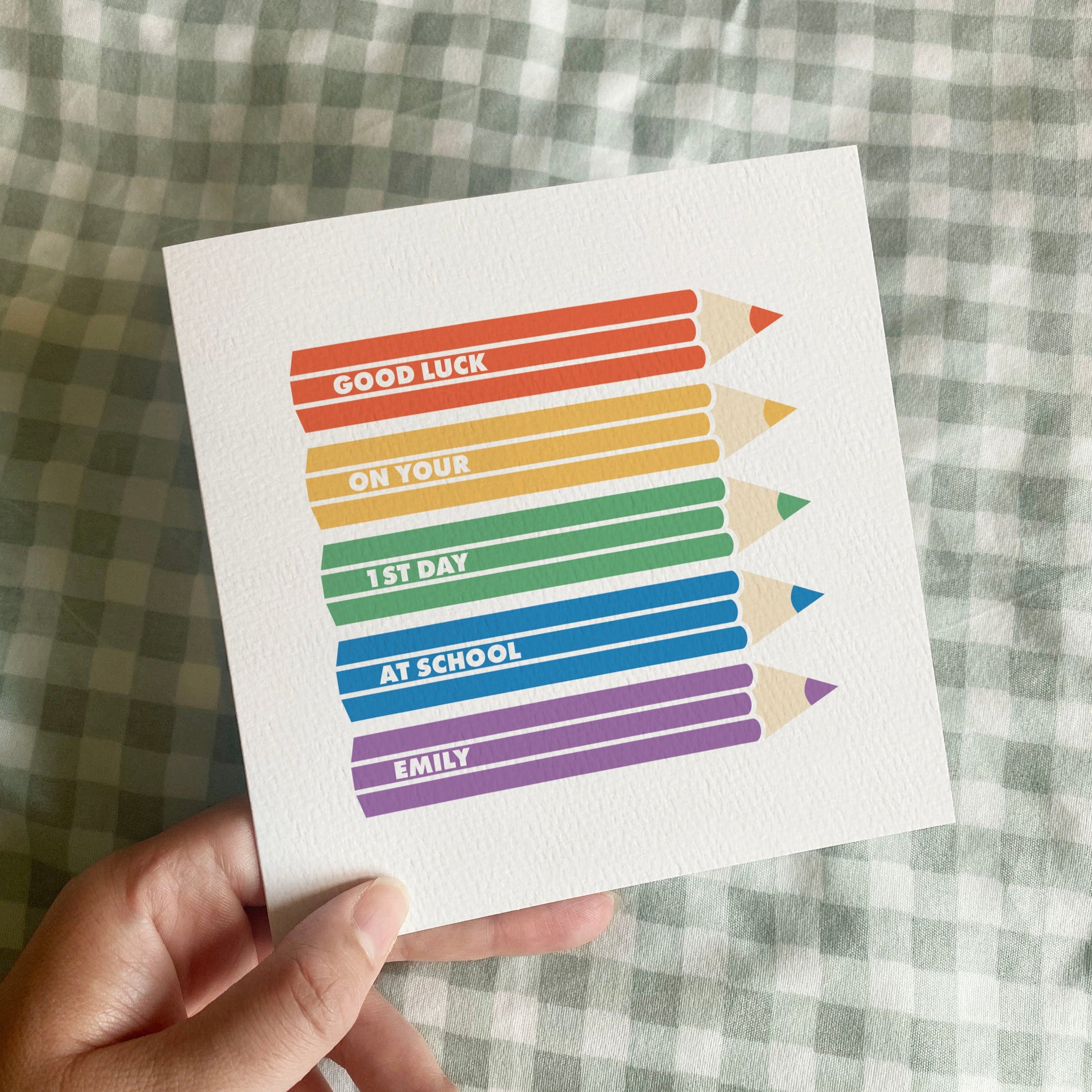 Hand holding a card with rainbow coloured pencils and text on a checkered fabric background