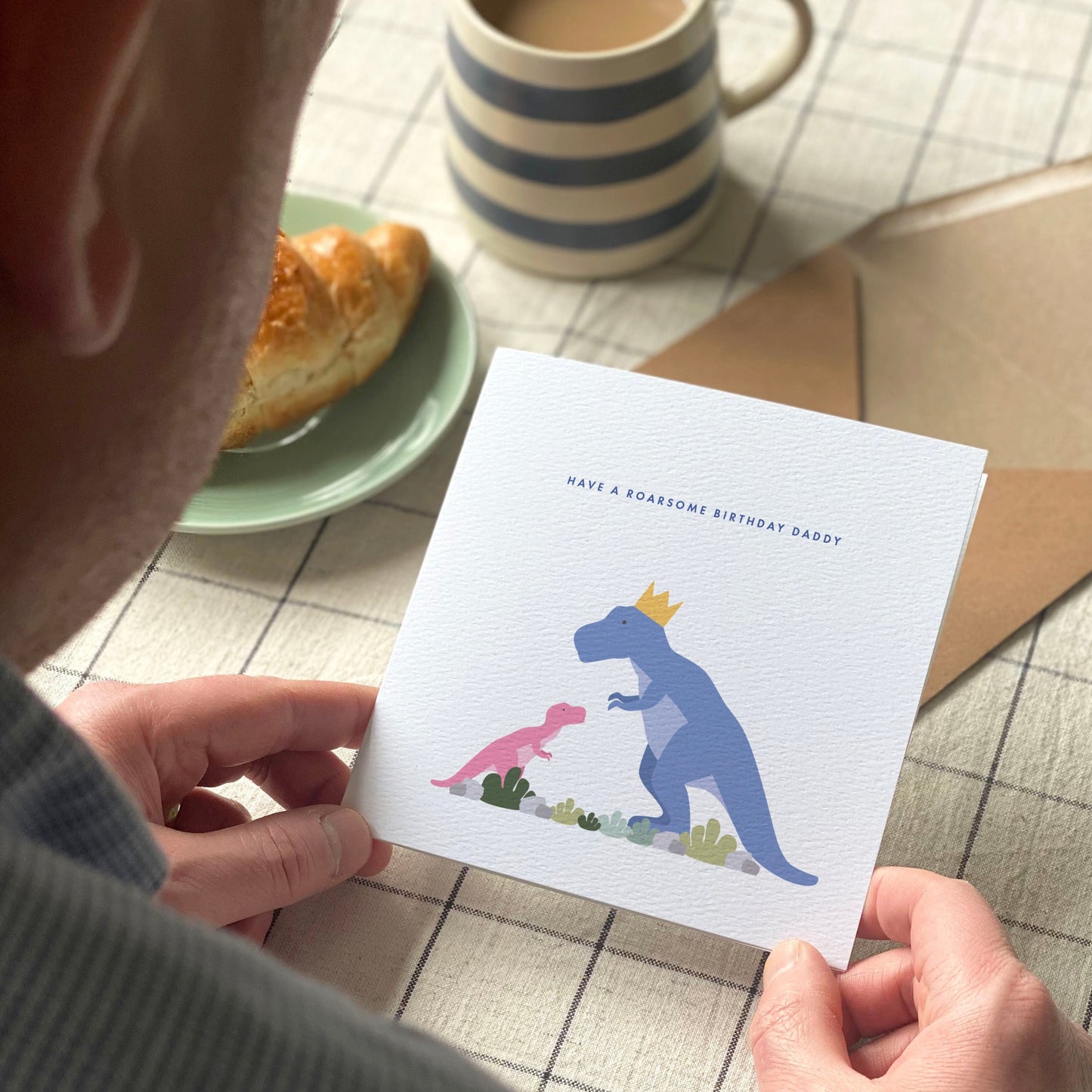 Man holding a card with dinosaur illustration on a table with a mug and pastry.