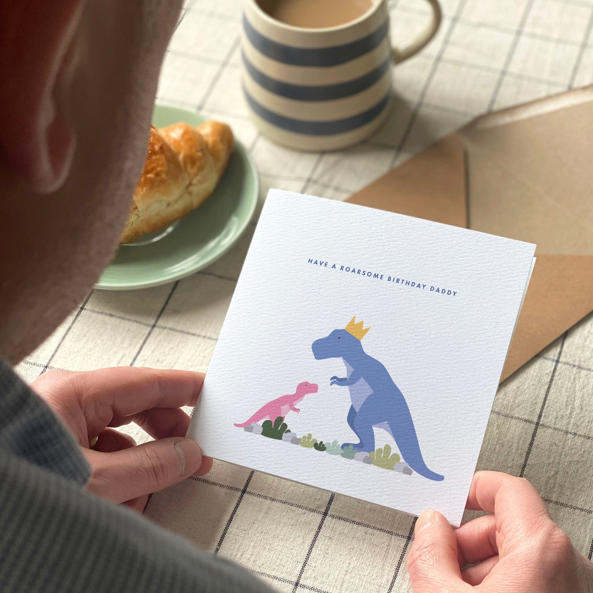 Man holding a card with dinosaur illustration on a table with a mug and pastry.