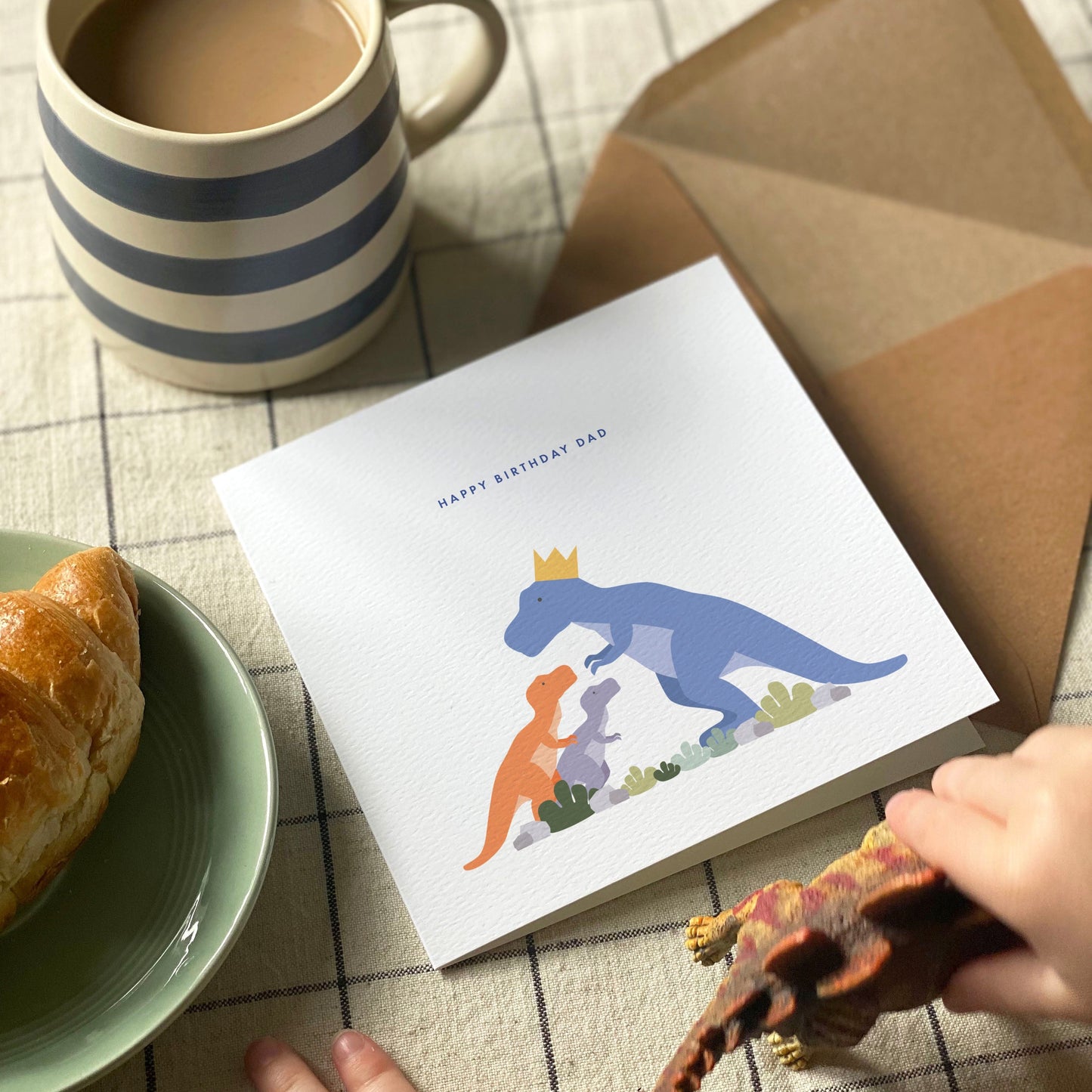 Dinosaur themed birthday card with a mug, croissant, and stegosaurus toy on a checkered tablecloth.