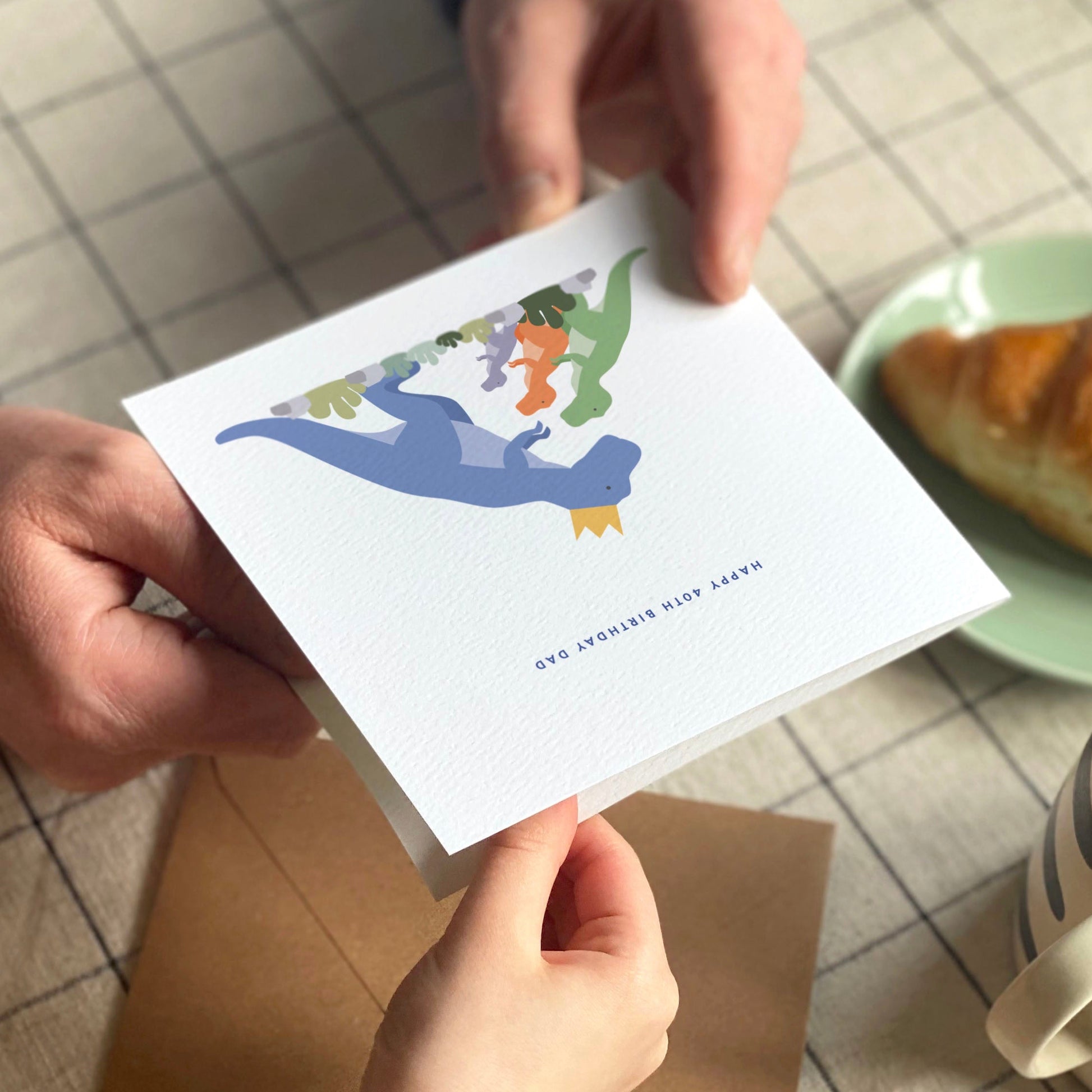 Father and child holding a colourful dinosaur birthday card with a croissant and coffee cup on a grid table cloth.