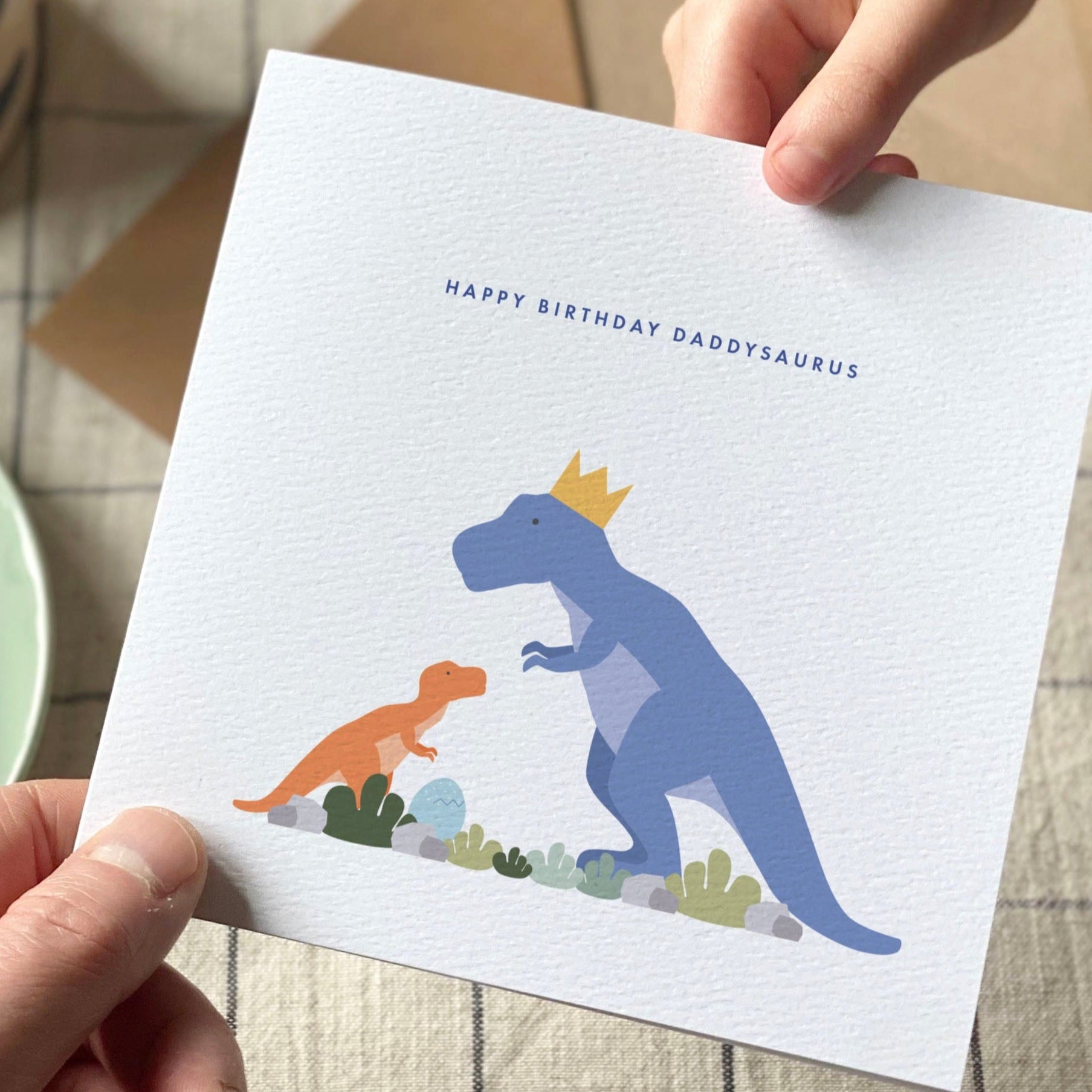 Person holding a birthday card with dinosaur illustration next to a cup of coffee and croissant on a checkered tablecloth.
