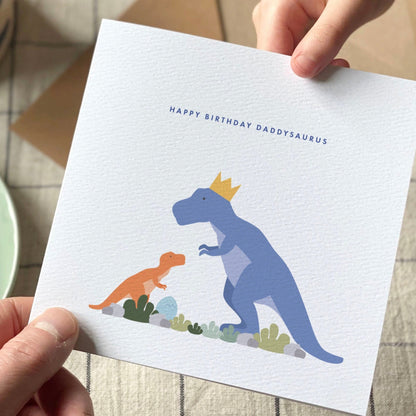Person holding a birthday card with dinosaur illustration next to a cup of coffee and croissant on a checkered tablecloth.