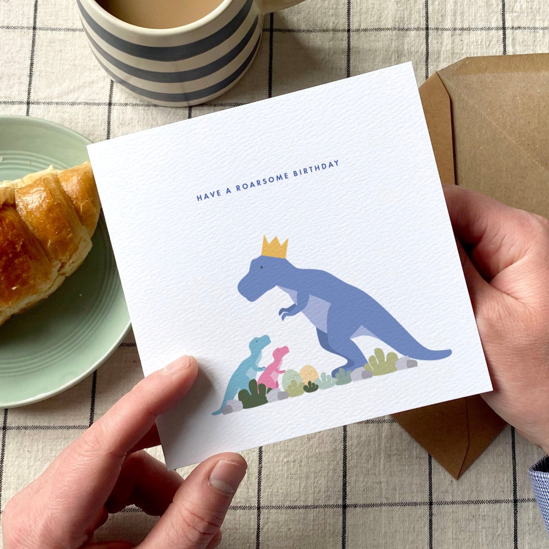 Person holding a card with a dinosaur illustration on a table with a croissant and coffee.